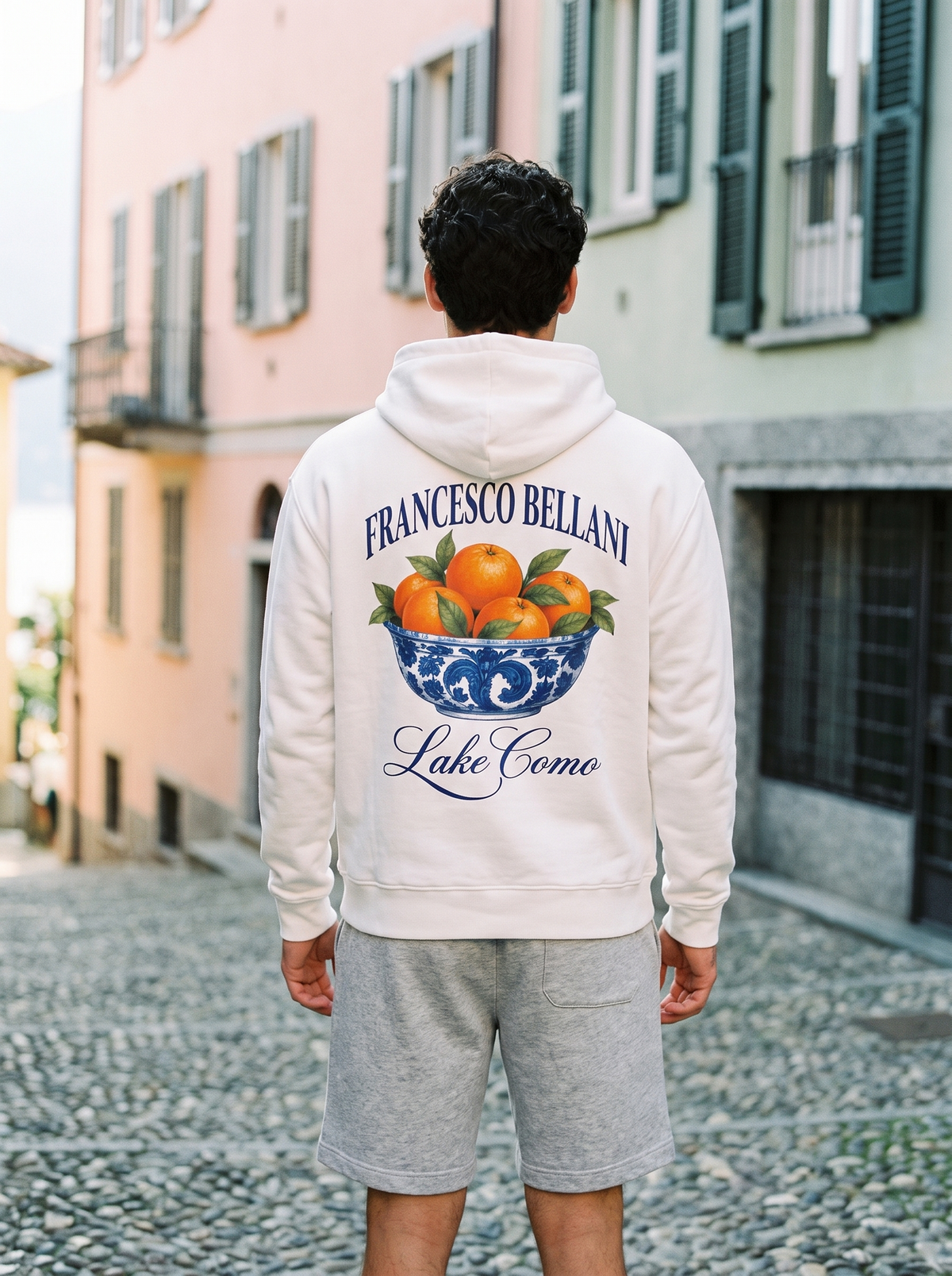 Man wearing the white Moltrasio Printed Hoodie with grey jersey shorts on a cobbled street in Como